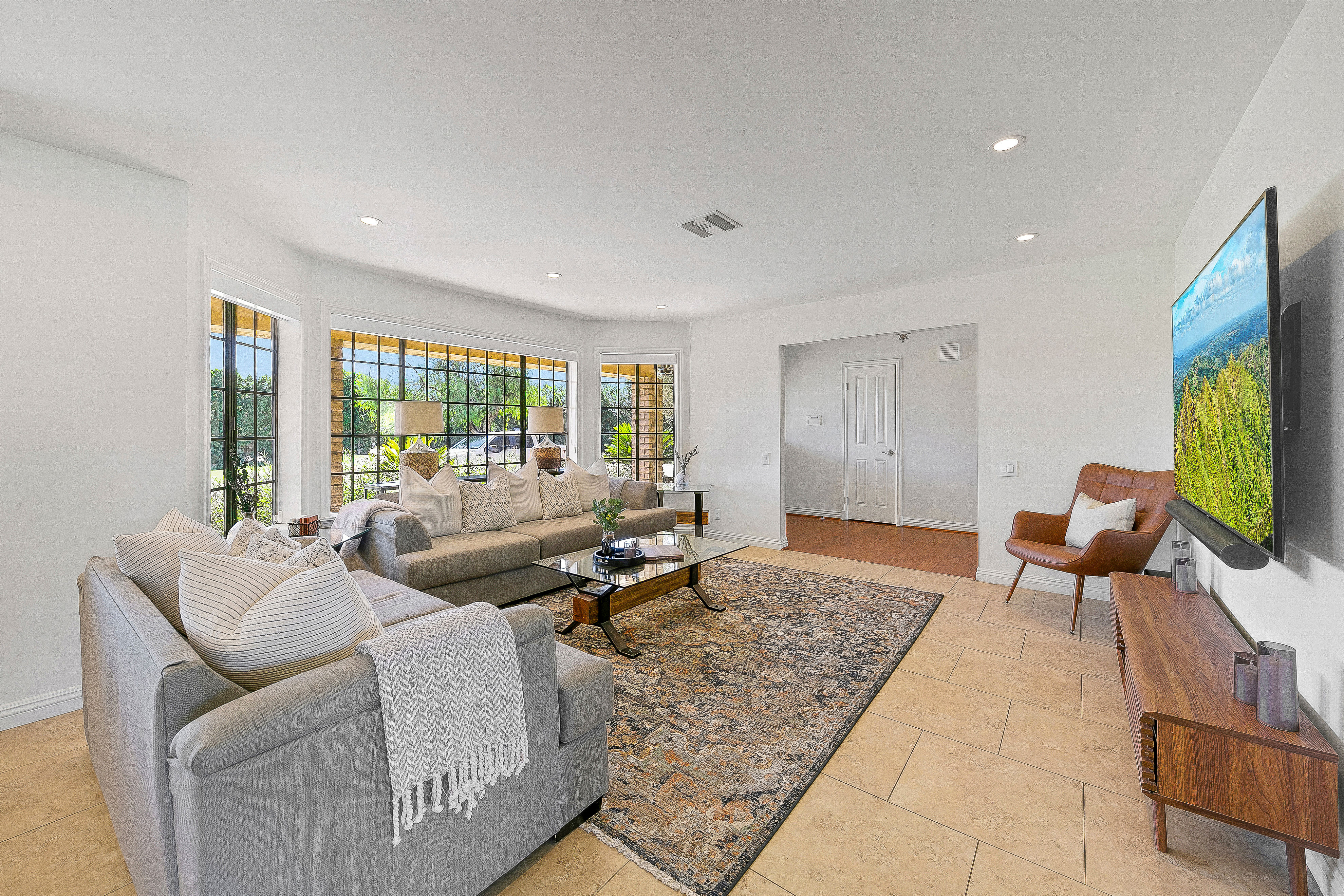 Sunlit modern living room with neutral sofas and throw pillows, large grid bay windows overlooking greenery, glass coffee table on a patterned area rug, wall-mounted TV and leather accent chair.