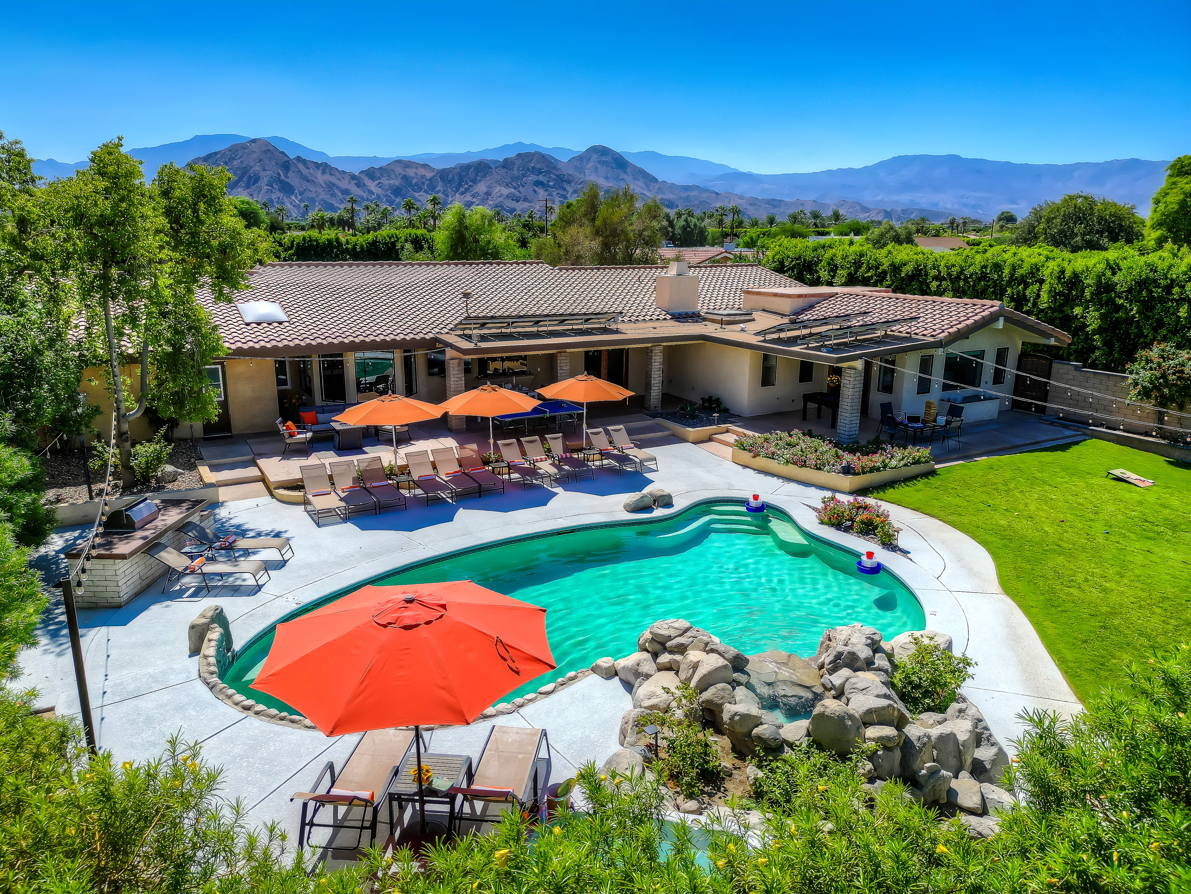 Aerial view of a backyard pool with rock waterfall, orange umbrellas and lounge chairs at a tile‑roof home, green lawn and desert mountain backdrop