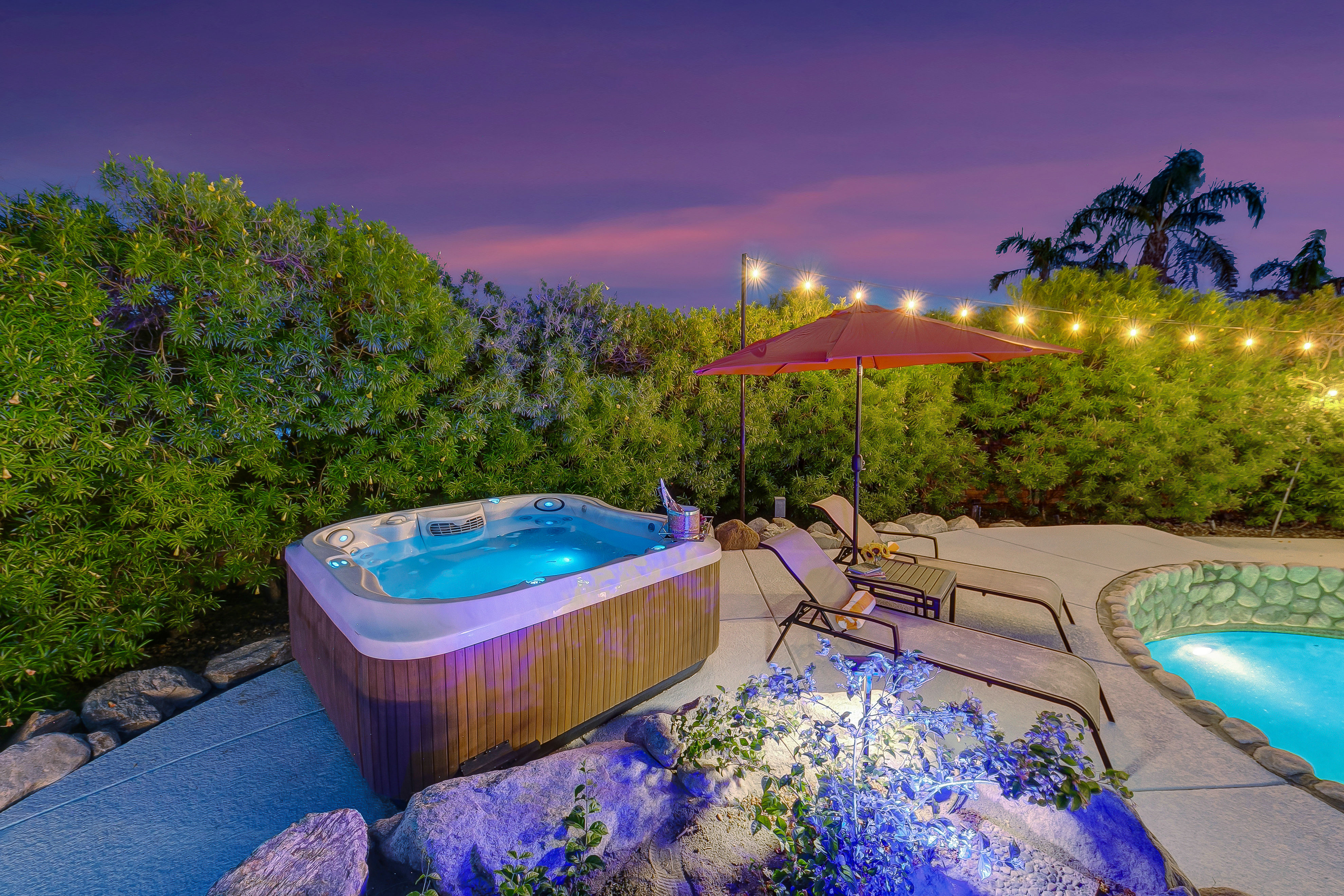 Backyard patio at dusk with an illuminated hot tub beside a glowing swimming pool, two lounge chairs under a red umbrella, string lights and lush tropical hedges beneath a purple sky.