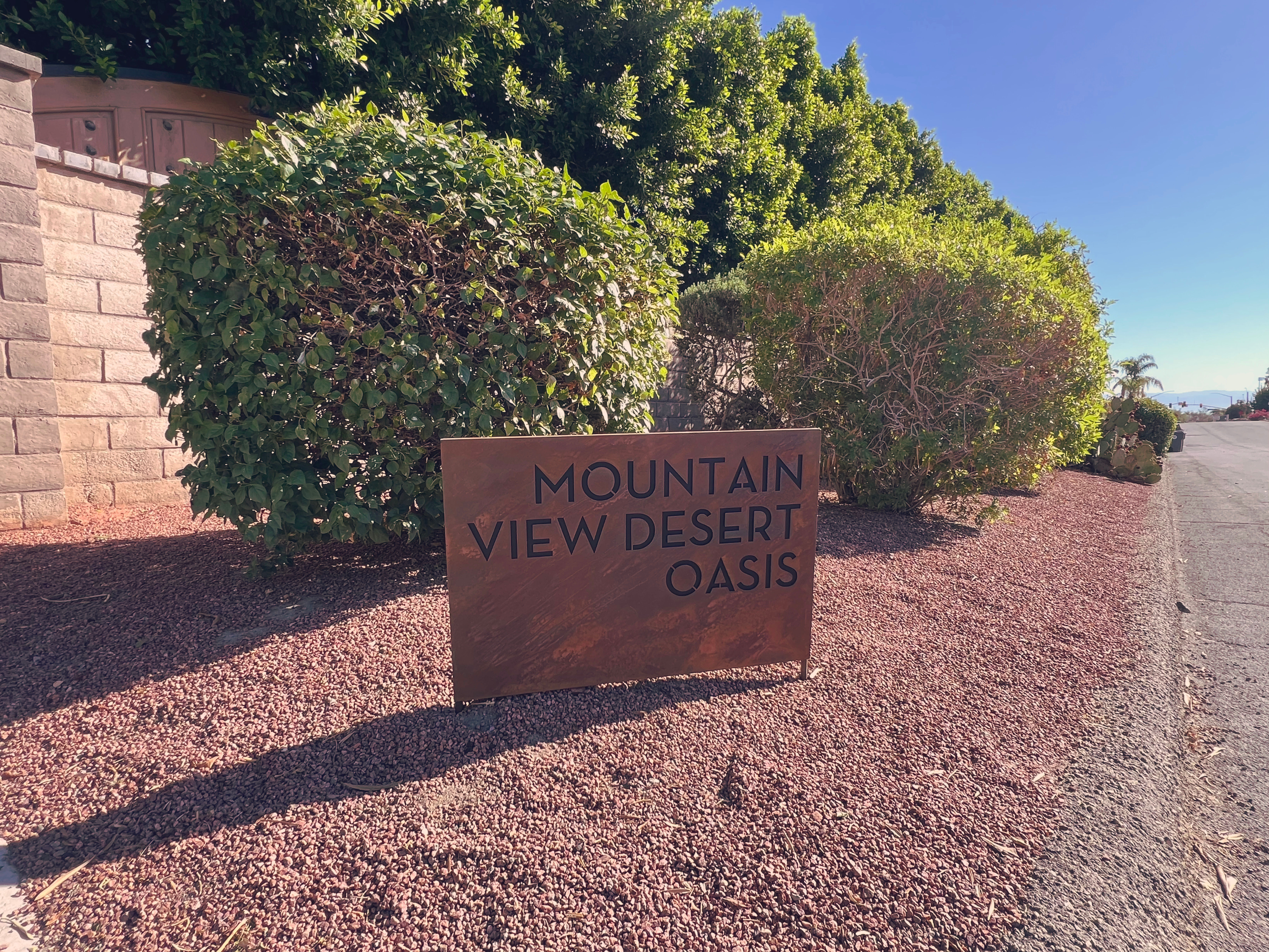 Rust-colored metal community sign in a desert streetscape, set on red gravel among trimmed green shrubs with a clear blue sky and distant mountain view along a quiet residential road.