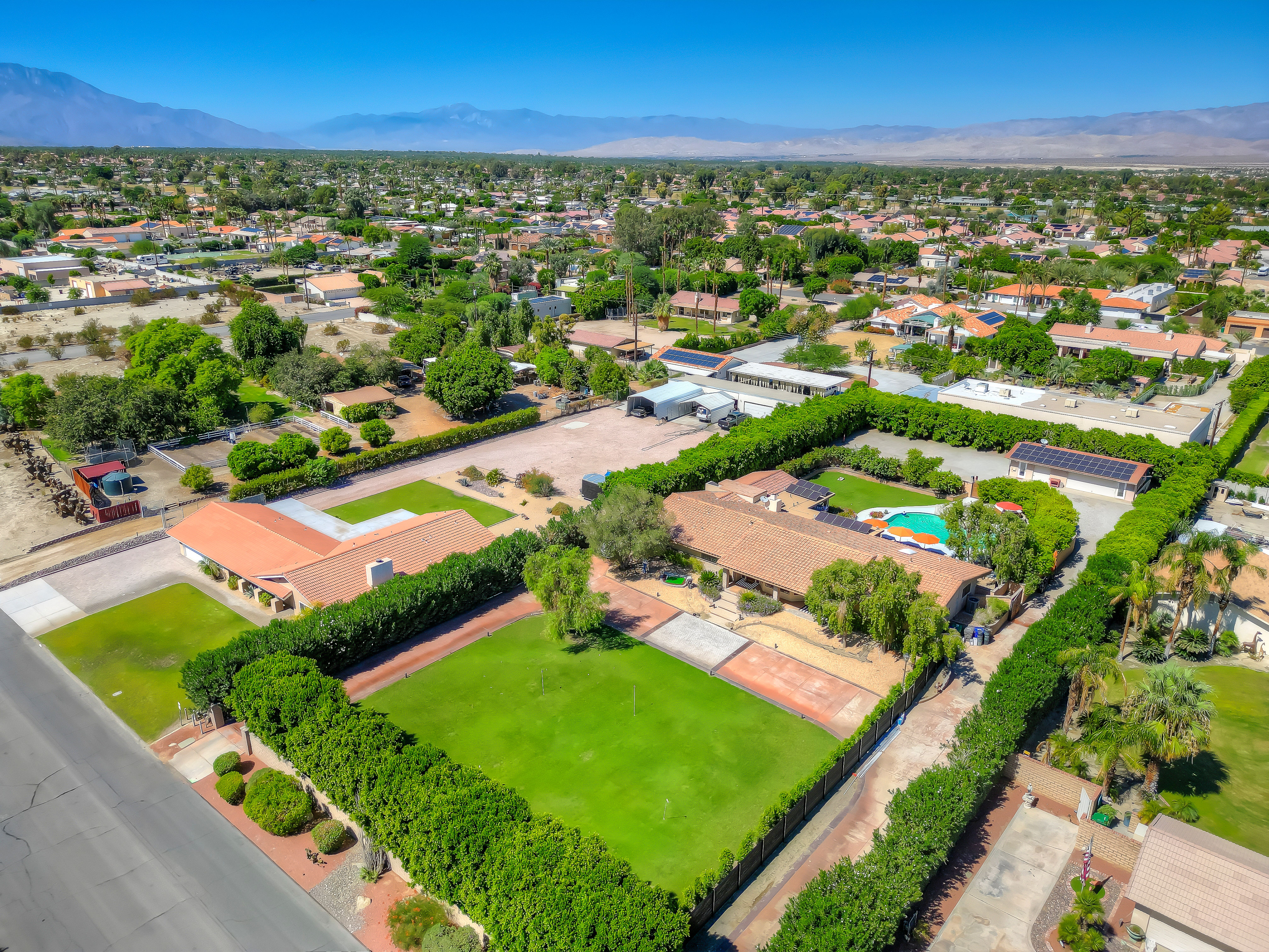 Aerial view of a sunlit suburban desert neighborhood with single-story ranch homes, lush hedged lawns, a backyard swimming pool and palm trees, and mountains on the distant horizon.