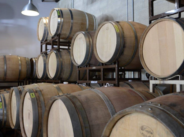 Rows of oak wine barrels stacked on metal racks in a bright winery barrel room, used for aging wine.