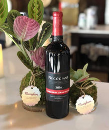 2016 Cabernet Sauvignon bottle as a table centerpiece with moss ball planters and pink-green tropical leaves in an indoor setting