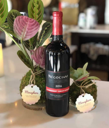 2016 Cabernet Sauvignon bottle as a table centerpiece with moss ball planters and pink-green tropical leaves in an indoor setting