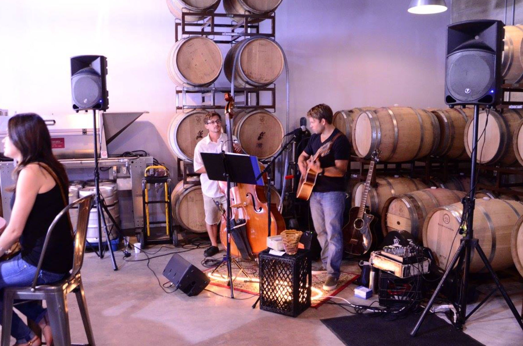 Acoustic duo performing in a winery barrel room among stacked oak wine barrels — guitarist and upright bass with microphones, speakers, and a few patrons at metal tables for an intimate live music tasting.