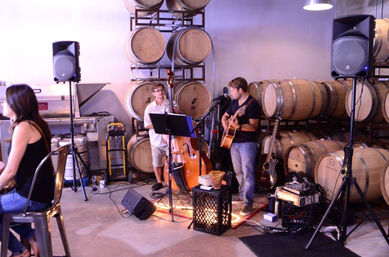 Acoustic duo performing in a winery barrel room among stacked oak wine barrels — guitarist and upright bass with microphones, speakers, and a few patrons at metal tables for an intimate live music tasting.