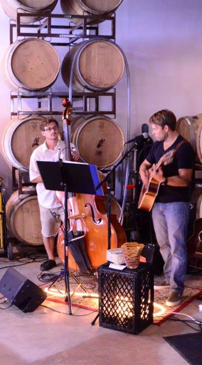 Acoustic duo performing in a winery barrel room among stacked oak wine barrels — guitarist and upright bass with microphones, speakers, and a few patrons at metal tables for an intimate live music tasting.