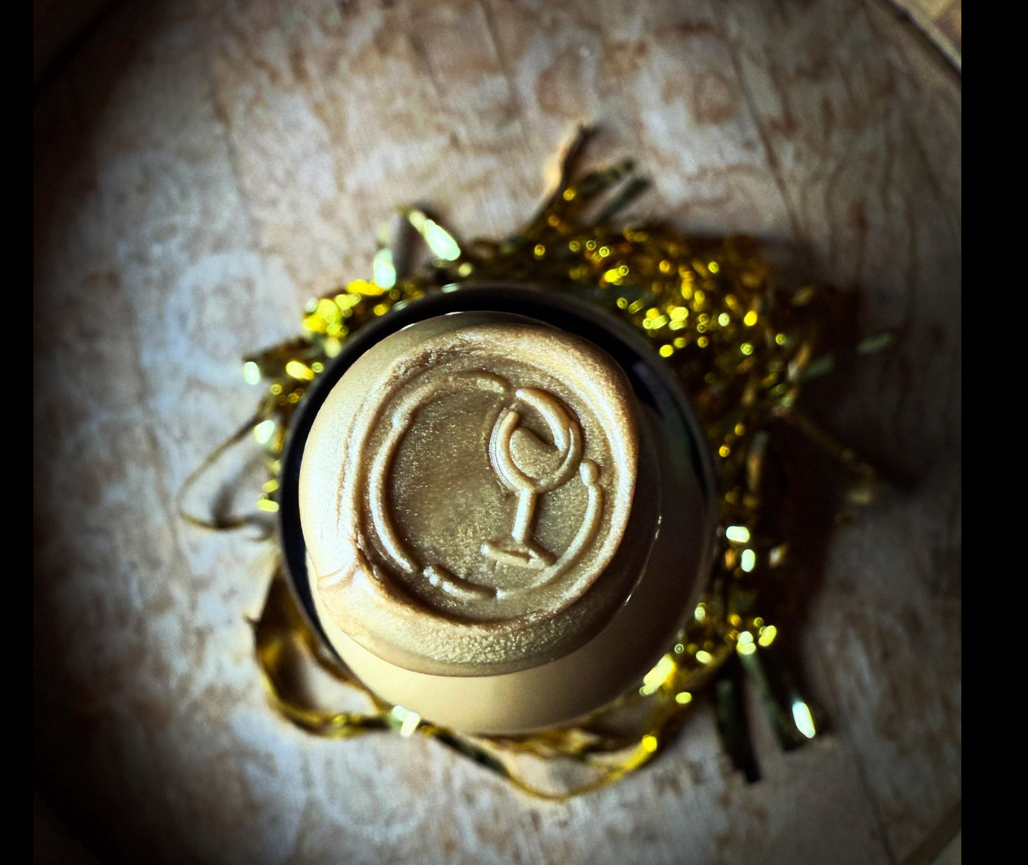Top-down close-up of a bottle top sealed with metallic gold wax embossed with a wine-glass icon, nestled in gold tinsel on a wooden surface