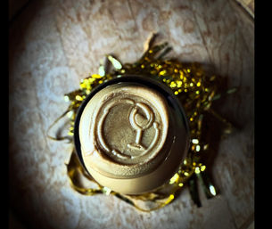 Top-down close-up of a bottle top sealed with metallic gold wax embossed with a wine-glass icon, nestled in gold tinsel on a wooden surface