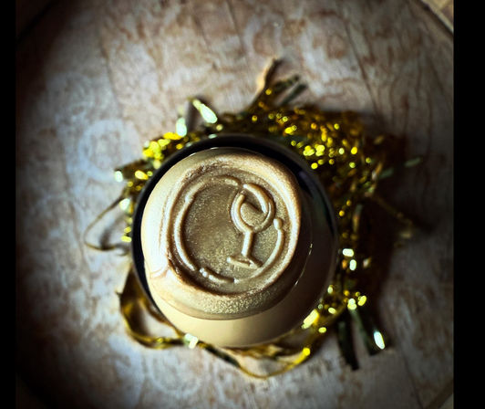 Top-down close-up of a bottle top sealed with metallic gold wax embossed with a wine-glass icon, nestled in gold tinsel on a wooden surface