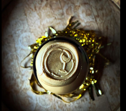 Top-down close-up of a bottle top sealed with metallic gold wax embossed with a wine-glass icon, nestled in gold tinsel on a wooden surface