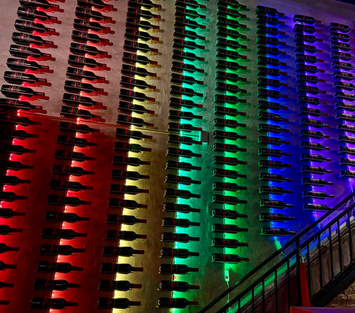 Wall of horizontally mounted wine bottles in neat rows, backlit by red, yellow, green and blue LED lights forming a rainbow gradient in a dim bar or restaurant interior.