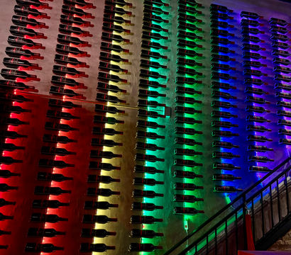 Wall of horizontally mounted wine bottles in neat rows, backlit by red, yellow, green and blue LED lights forming a rainbow gradient in a dim bar or restaurant interior.