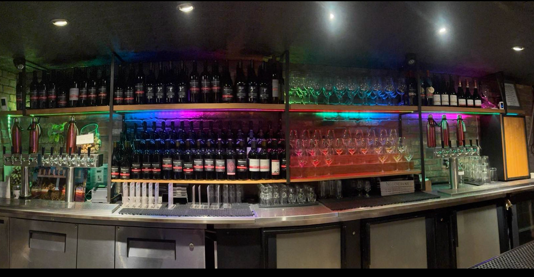 Indoor wine bar counter with shelves of wine bottles and rows of hanging wine glasses, colorful LED backlighting, multiple beer taps and stainless-steel counter with refrigerated cabinets