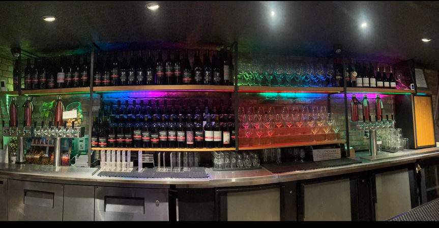 Indoor wine bar counter with shelves of wine bottles and rows of hanging wine glasses, colorful LED backlighting, multiple beer taps and stainless-steel counter with refrigerated cabinets