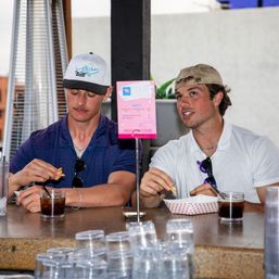 Two men in baseball caps at an outdoor rooftop bar counter on an urban patio, snacking on fries from a paper basket and sipping dark sodas with sunglasses clipped to their shirts and a pink menu sign on the counter.
