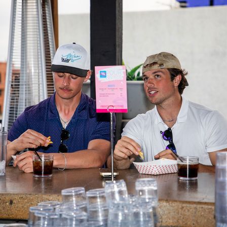 Two men in baseball caps at an outdoor rooftop bar counter on an urban patio, snacking on fries from a paper basket and sipping dark sodas with sunglasses clipped to their shirts and a pink menu sign on the counter.