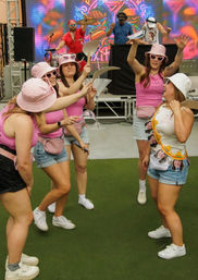 Group of women in matching pink tank tops, bucket hats and heart-shaped sunglasses dancing and waving paper props on green turf in front of a colorful LED DJ stage at an outdoor daytime music event