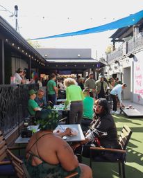 Sunlit urban outdoor patio bar courtyard with a lively crowd in green, people seated at tables, mingling and playing cornhole under string lights and a blue shade sail.