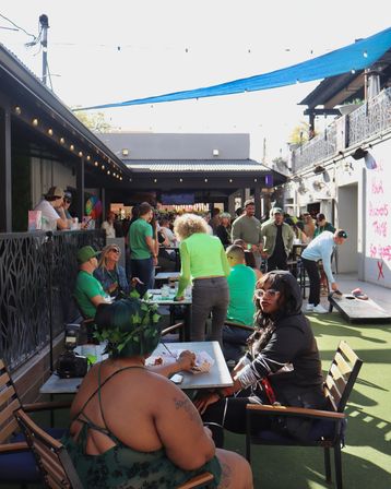 Sunlit urban outdoor patio bar courtyard with a lively crowd in green, people seated at tables, mingling and playing cornhole under string lights and a blue shade sail.