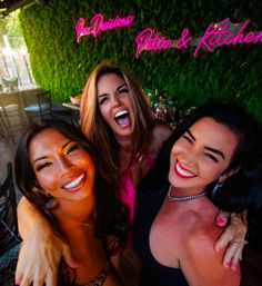 Three friends laughing in a selfie on a lively outdoor patio with a green plant wall and bright pink neon "Patio & Kitchen" sign