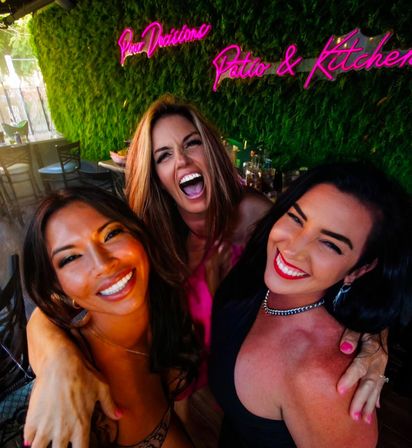 Three friends laughing in a selfie on a lively outdoor patio with a green plant wall and bright pink neon "Patio & Kitchen" sign