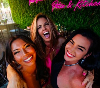 Three women laughing and hugging for a selfie on a lively outdoor patio bar with a pink neon sign and green living wall