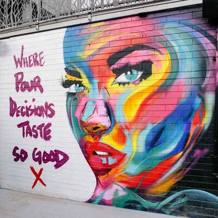 Vibrant street-art mural of a multicolored stylized female face on a brick wall, with bold purple graffiti text reading "WHERE POOR DECISIONS TASTE SO GOOD", colorful urban portrait.