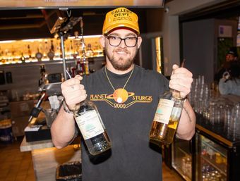 Smiling bartender in a yellow cap and glasses holding two large clear and amber spirit bottles in a brightly lit bar with rows of bottles and glassware