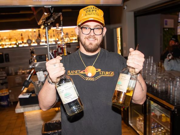 Smiling bartender in a yellow cap and glasses holding two large clear and amber spirit bottles in a brightly lit bar with rows of bottles and glassware