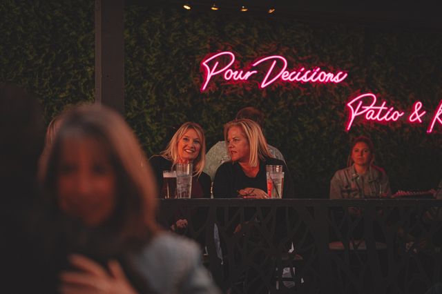 Two women chatting at an outdoor bar patio at night with drinks on the railing, neon pink sign glowing on a leafy wall and other patrons in the background.