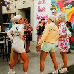 Group of friends laughing and dancing on an outdoor patio beside a colorful urban mural, women in summer outfits and sneakers at a casual street‑art gathering