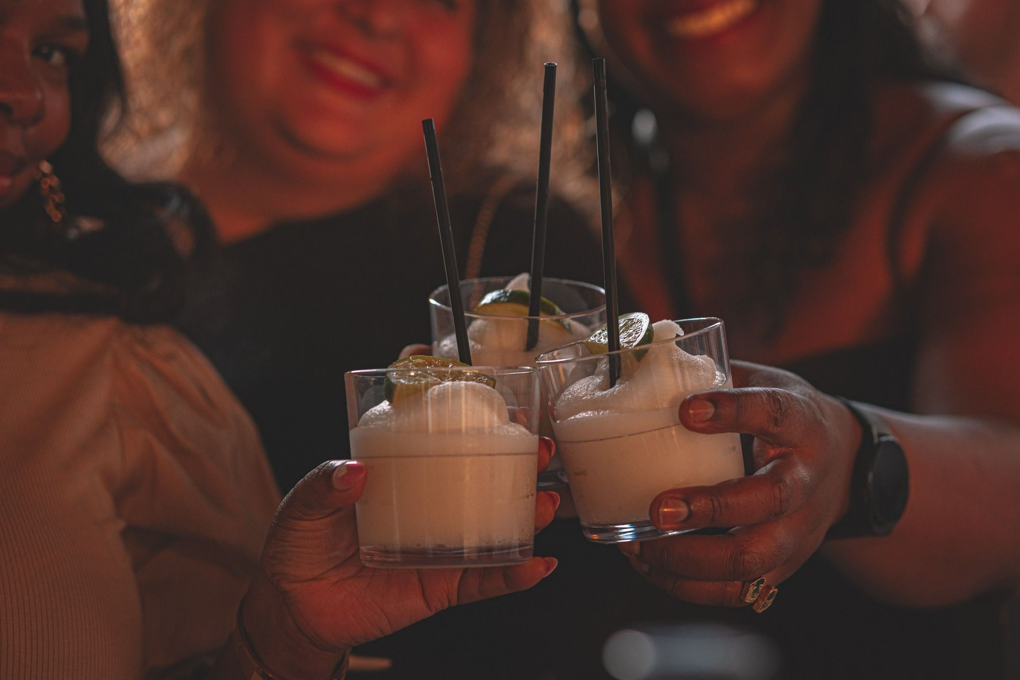 Three frothy frozen lime cocktails with black straws being toasted by people in a dimly lit bar, lively nightlife vibe