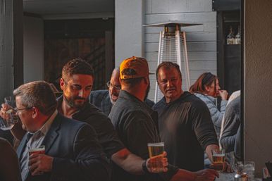Casual group of men socializing on a busy bar patio at sunset, holding pints of beer near a tall outdoor heater