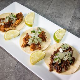 Three street-style corn tacos on a white platter—two with shredded chicken and one with seasoned beef—topped with chopped onion and cilantro and served with lime wedges on a stone tabletop.