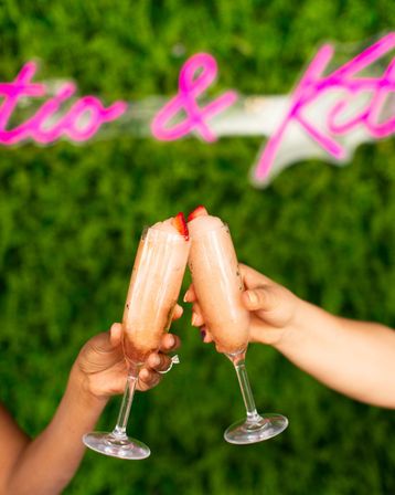 Two hands clinking pink strawberry frozen mimosas in champagne flutes with strawberry garnishes, set against a green hedge wall and blurred neon pink sign for an outdoor brunch celebration vibe.