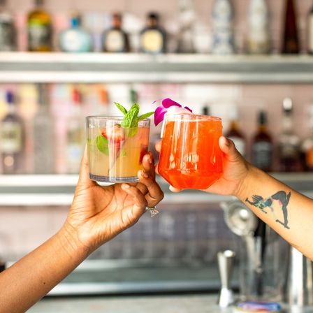 Two hands clinking colorful craft cocktails at a lively bar during happy hour — mint-garnished citrus drink and bright orange orchid-topped cocktail.