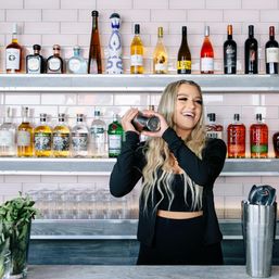 Cheerful bartender shaking a cocktail shaker at a modern bar with pink subway-tile backsplash, rows of liquor bottles on stainless shelves and stacked glassware