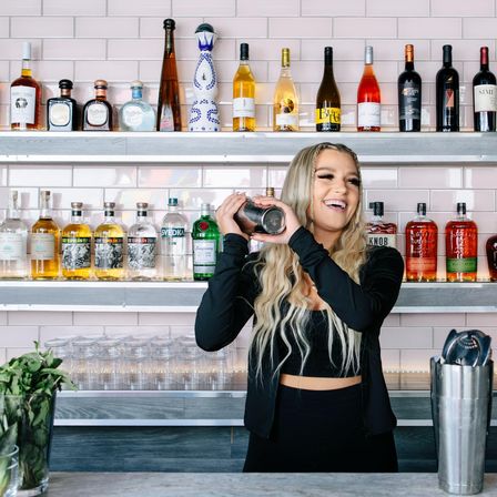 Cheerful bartender shaking a cocktail shaker at a modern bar with pink subway-tile backsplash, rows of liquor bottles on stainless shelves and stacked glassware