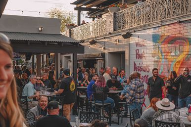 Busy urban outdoor patio at sunset with people socializing around high-top tables, string lights, rooftop terrace and a colorful graffiti mural.