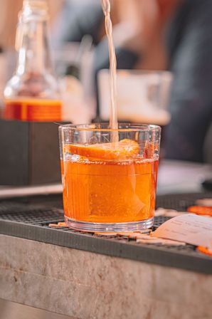 Amber cocktail being poured into a rocks glass with an orange slice garnish on a bar counter, close-up of a refreshing cocktail pour