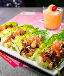 Grilled chicken lettuce wraps with diced tomato pico and balsamic drizzle on a white plate, photographed on a pink menu with a peach cocktail garnished with lemon and a cherry in a casual restaurant setting.