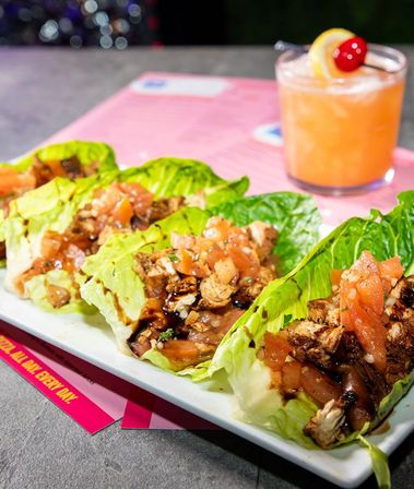 Grilled chicken lettuce wraps with diced tomato pico and balsamic drizzle on a white plate, photographed on a pink menu with a peach cocktail garnished with lemon and a cherry in a casual restaurant setting.