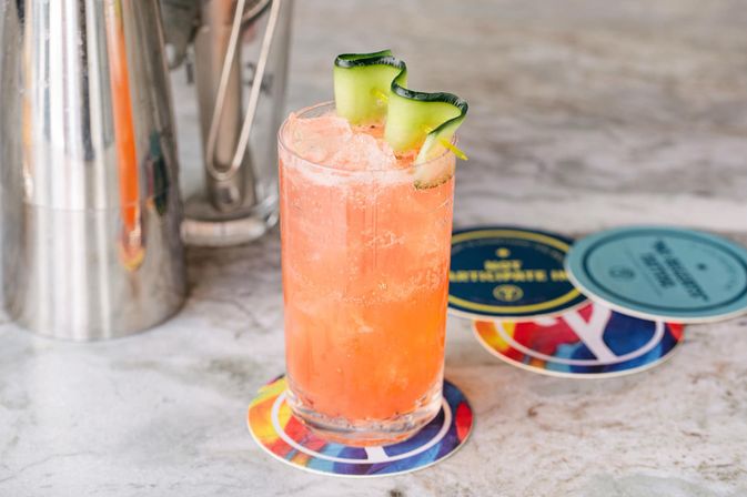 Bright pink-orange craft cocktail with crushed ice and a curled cucumber ribbon garnish in a highball glass, sitting on a colorful coaster on a marble bar countertop beside a stainless-steel cocktail shaker.