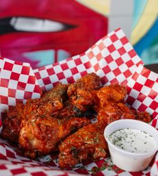 Close-up of saucy buffalo chicken wings on red-and-white checkered paper, sprinkled with herbs and served with a cup of ranch dip in front of a colorful mural backdrop.