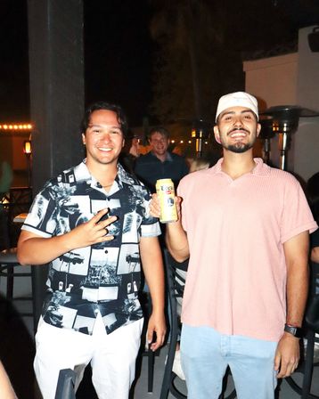 Two friends smiling on a lively outdoor patio at night — one in a black-and-white patterned shirt flashing a peace sign, the other in a pink shirt and white cap holding a canned beer with string lights and patio heaters glowing behind them.