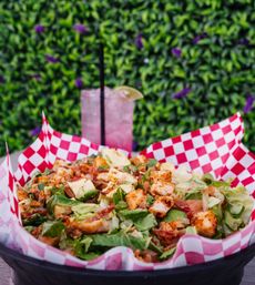 Grilled chicken salad with chopped romaine, avocado, crispy bacon and seasoned chicken bites served in a red-and-white checkered paper-lined basket on an outdoor patio, with a pink iced lemonade garnished with lime in the background.