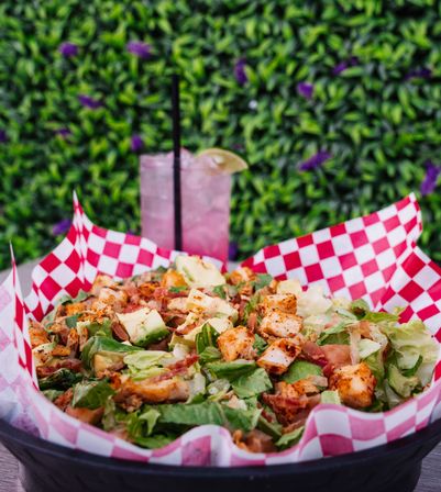 Grilled chicken salad with chopped romaine, avocado, crispy bacon and seasoned chicken bites served in a red-and-white checkered paper-lined basket on an outdoor patio, with a pink iced lemonade garnished with lime in the background.