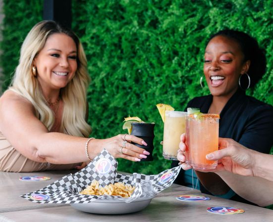 Two friends toasting colorful cocktails and a small black cup over a table with fries on checkered paper at an outdoor patio with a lush green hedge—bright, brunch-style scene.