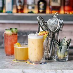 Three colorful craft cocktails on a marble bar counter: a tall frothy tropical iced drink garnished with pineapple wedge and cinnamon, a small citrus cocktail, and a pink cucumber‑garnished spritz, with metal cocktail strainers and pineapple leaves in the background.